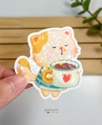 A die-cut waterproof vinyl sticker of an adorable cream and orange striped cat happily holding a giant mint green mug with a red heart on it. The mug is filled with a warm drink, and the sticker is finished with a vibrant star holographic sparkle overlay.