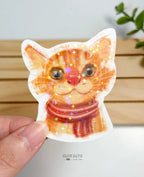 A die-cut waterproof vinyl sticker showing a close-up portrait illustration of an adorable orange tabby cat wearing a red and brown striped scarf. The entire sticker is finished with a vibrant star holographic sparkle overlay.