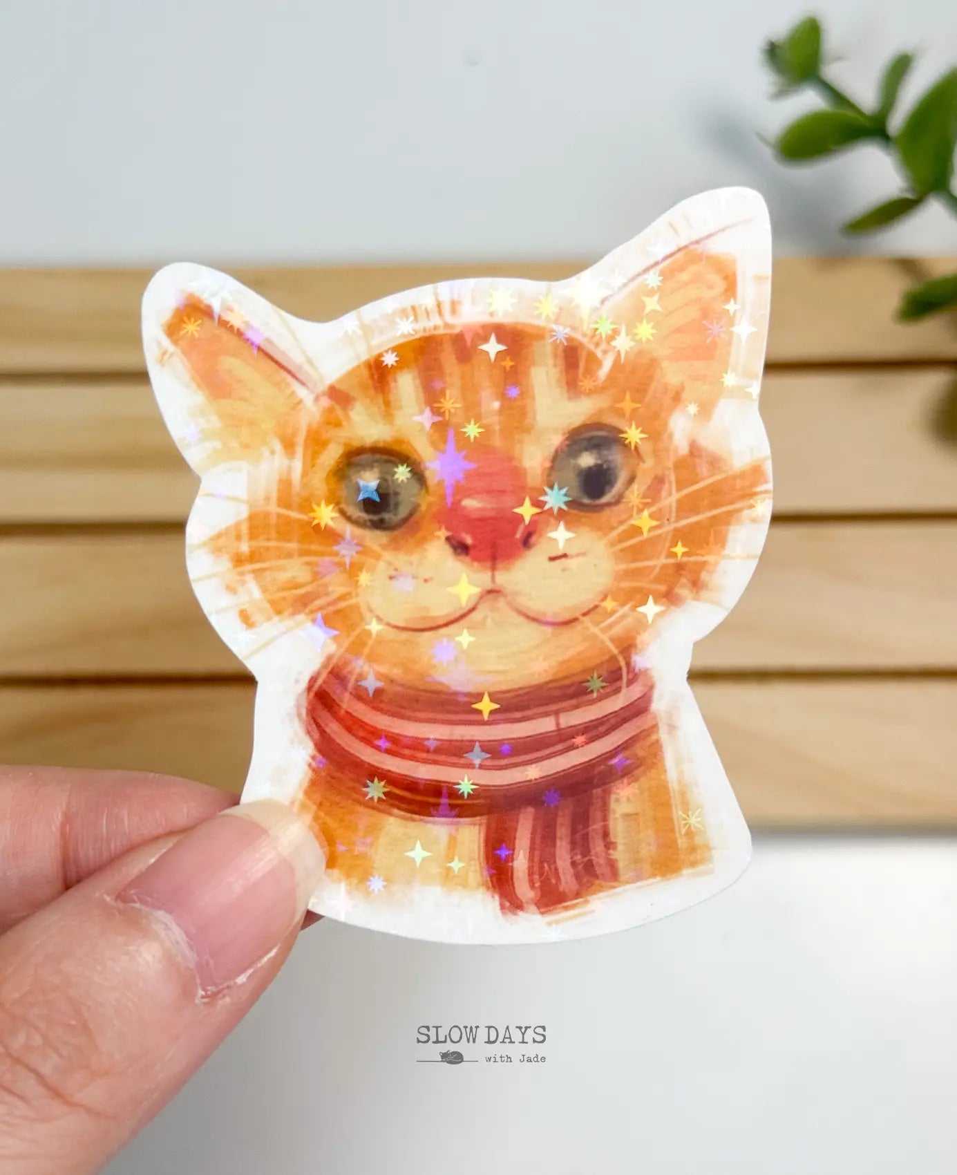 A die-cut waterproof vinyl sticker showing a close-up portrait illustration of an adorable orange tabby cat wearing a red and brown striped scarf. The entire sticker is finished with a vibrant star holographic sparkle overlay.