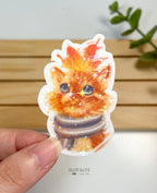A die-cut waterproof vinyl sticker showing a close-up portrait illustration of a cute orange tabby cat wearing a striped winter scarf with a large orange autumn maple leaf resting on its head, laminated in a star holographic sparkle finish.