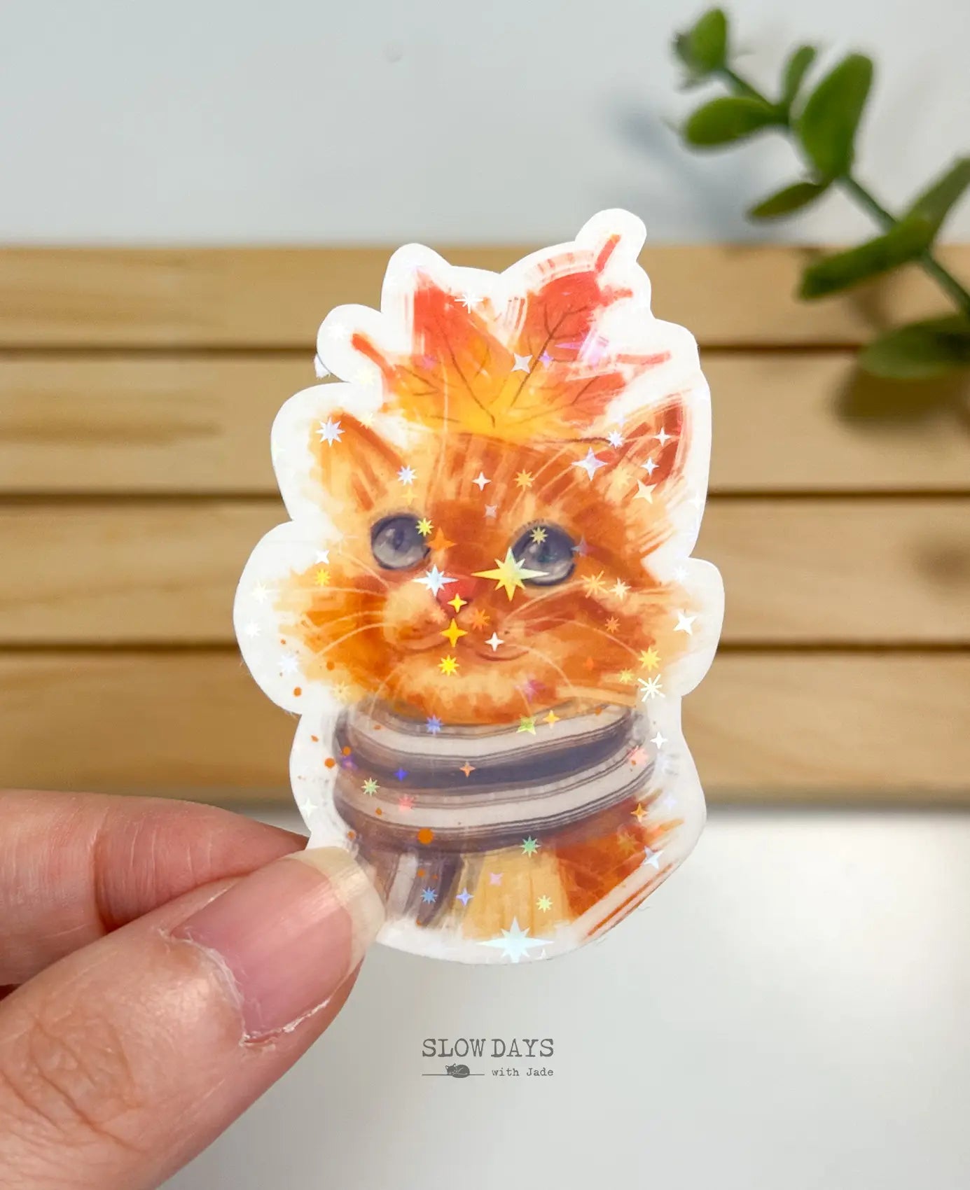 A die-cut waterproof vinyl sticker showing a close-up portrait illustration of a cute orange tabby cat wearing a striped winter scarf with a large orange autumn maple leaf resting on its head, laminated in a star holographic sparkle finish.