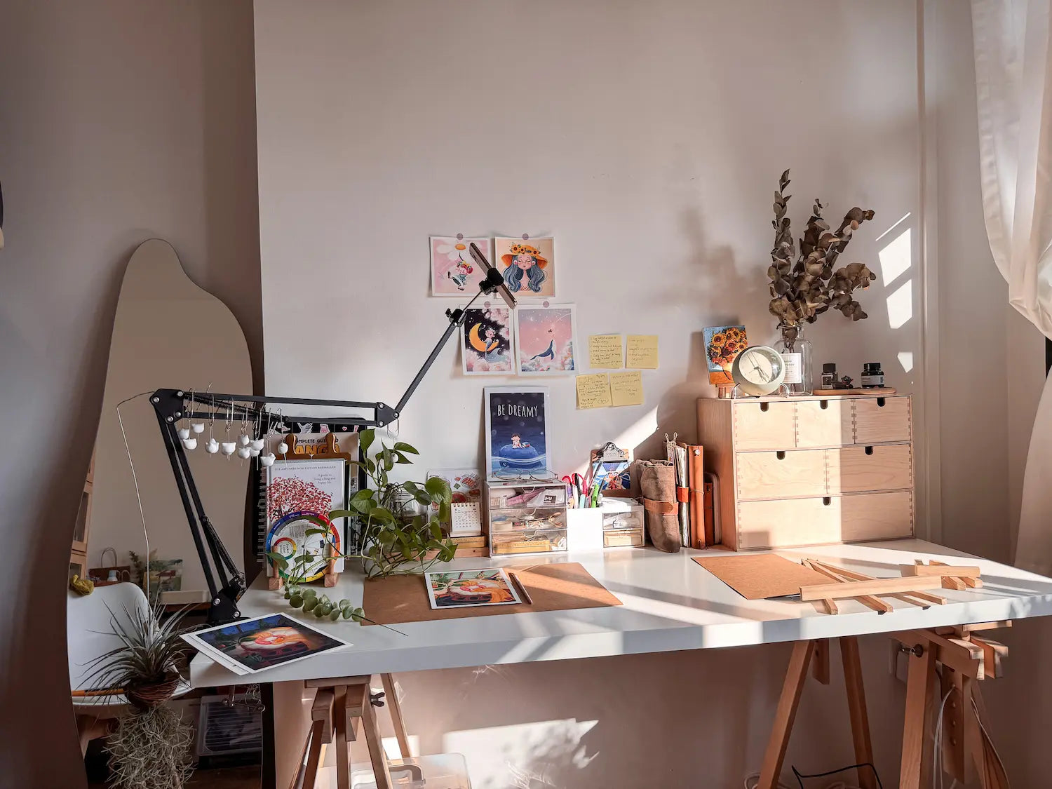 An image showcasing a cozy, sunlit artist's desk setup. The long white tabletop rests on wooden sawhorses, with bright sunlight streaming in from a window on the right casting long shadows. On the desk are brown kraft paper mats, art prints, a wooden drawer organizer topped with a clock and dried eucalyptus, a green pothos plant, and a jar of pens. Colorful art prints and yellow sticky notes are taped to the white wall behind it, and a wavy floor mirror stands to the far left.