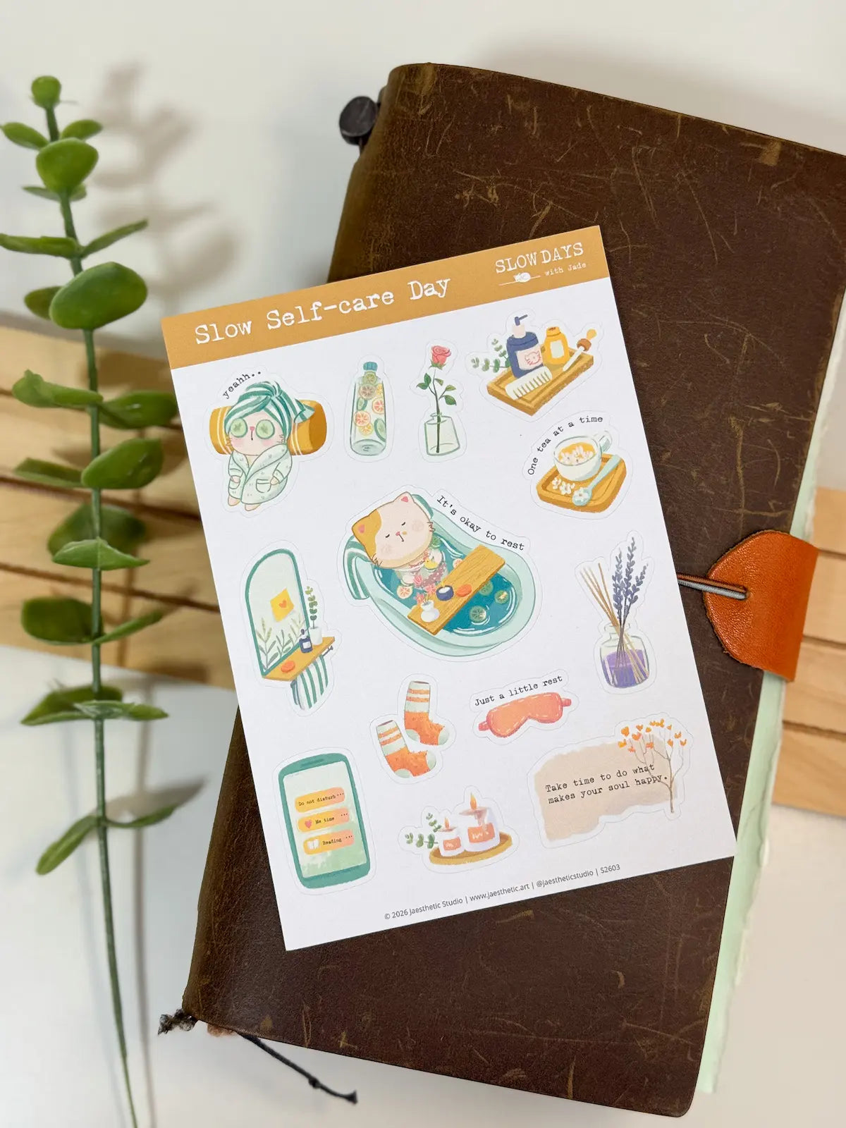 A full lifestyle flat lay photo of the "Slow Self-care Day" sticker sheet. The sheet is laid flat across a vintage brown leather notebook, with a faux green plant vine visible along the left edge, showing the complete collection of self-care and bath-themed cat stickers.