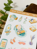 An angled close-up photo of a sticker sheet titled "Slow Self-care Day." The stickers feature cozy, wellness-themed illustrations, including a cute cat relaxing in a bubble bath, a cute cat character with cucumber eye slices in a bathrobe, striped towel, an eye mask, and a bottle of fruit-infused water.
