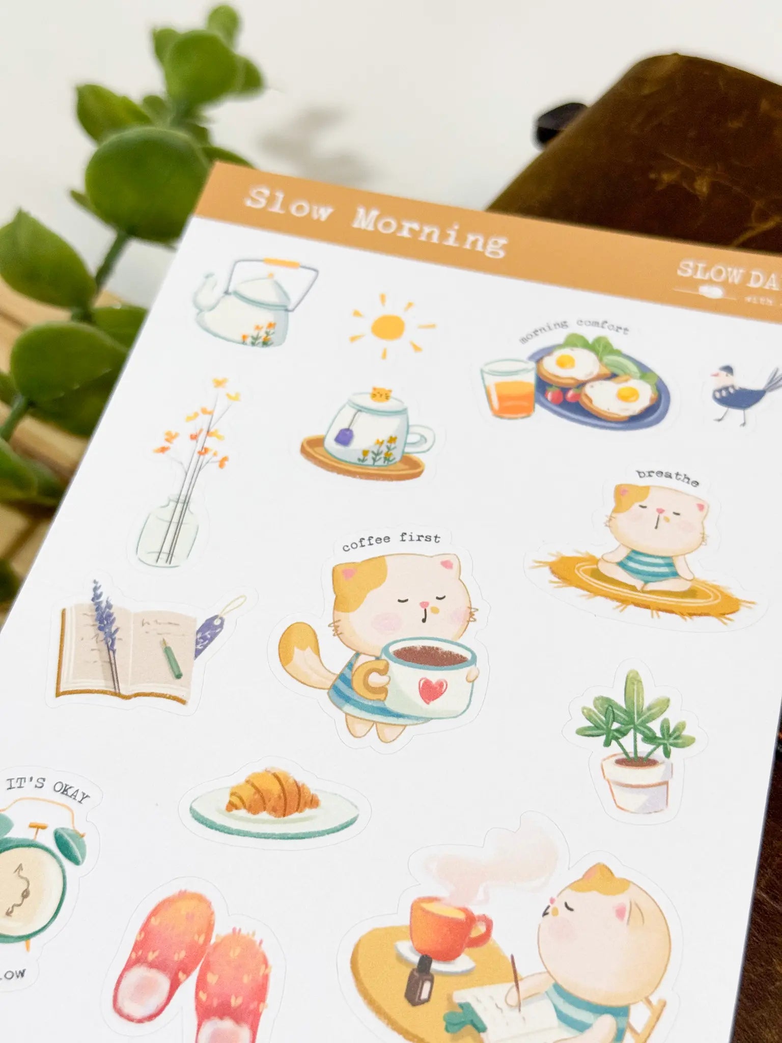 An angled close-up photo of a sticker sheet titled "Slow Morning." The warm-toned stickers include a cute cat happily holding a giant coffee mug with a red heart, a cat meditating on a straw mat with the word "breathe," a breakfast plate with eggs, a kettle, and fluffy orange slippers.