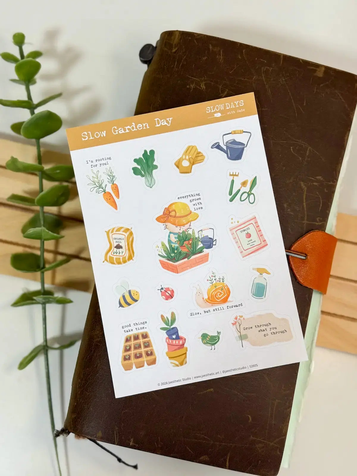 A full lifestyle flat lay photo of the "Slow Garden Day" sticker sheet positioned over a brown leather journal with a bright orange clasp. The shot captures the full sheet of gardening-themed cat and plant stickers, with a green stem framing the left side.