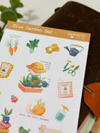 An angled close-up photo of a sticker sheet titled "Slow Garden Day." The cozy illustrations include a cat character in a sun hat tending to potted plants, smiling carrots with the text "I'm rooting for you!", gardening gloves, a watering can, a snail with plants growing on its shell, and a packet of tomato seeds.