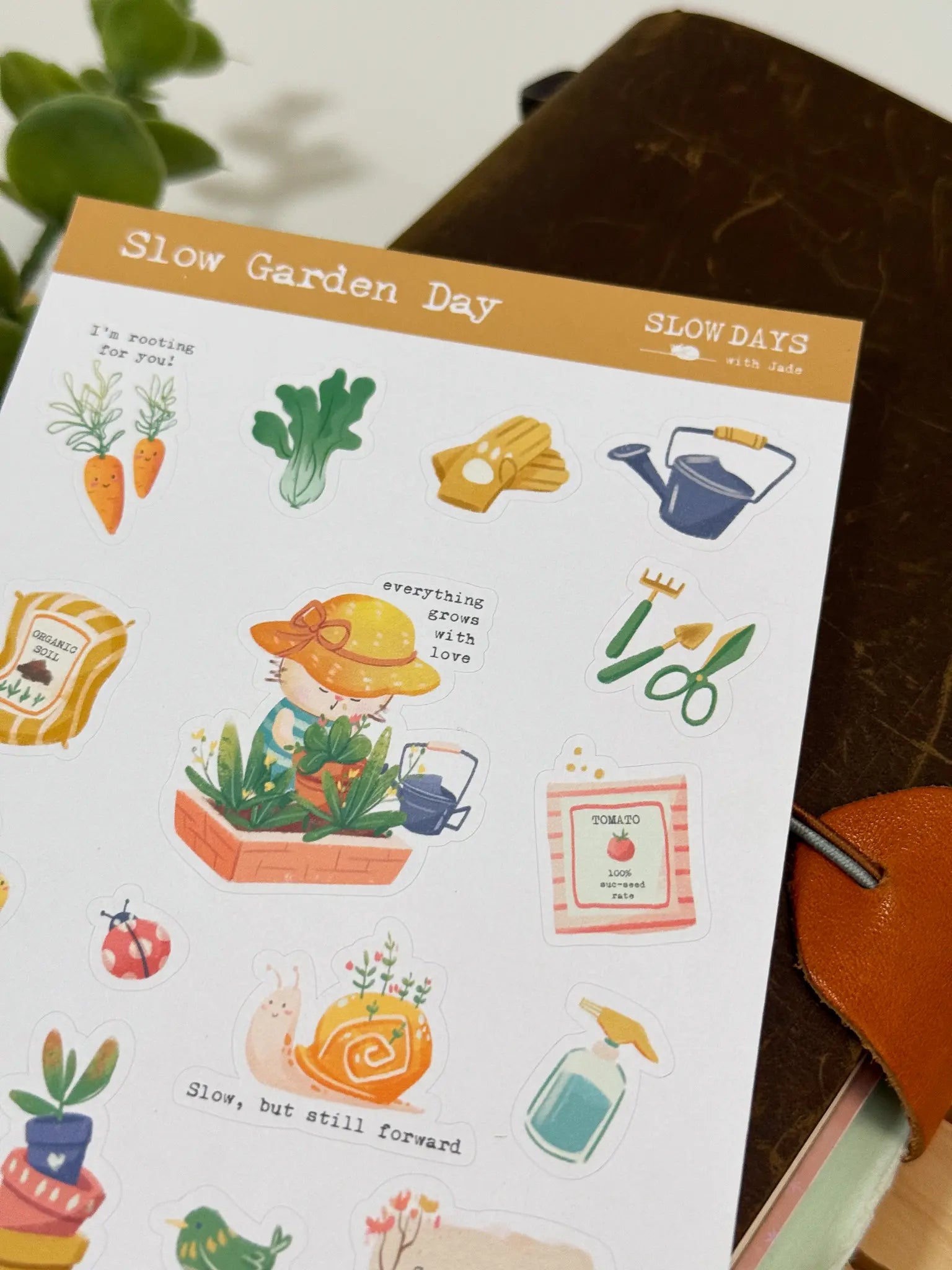 An angled close-up photo of a sticker sheet titled "Slow Garden Day." The cozy illustrations include a cat character in a sun hat tending to potted plants, smiling carrots with the text "I'm rooting for you!", gardening gloves, a watering can, a snail with plants growing on its shell, and a packet of tomato seeds.