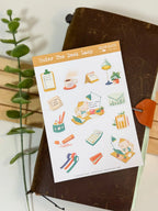 A full lifestyle flat lay photo of the "Under The Desk Lamp" sticker sheet resting on a dark brown leather traveler's notebook. The entire sheet of desk and stationery-themed cat stickers is visible, accented by a green plant stem on the left side.