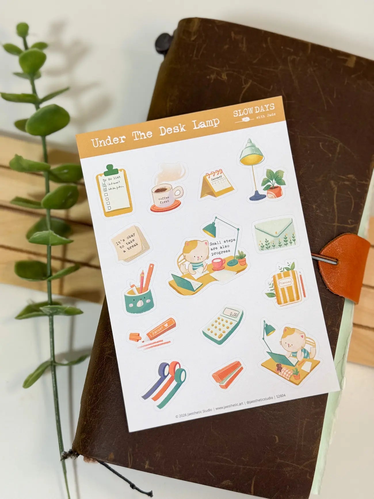 A full lifestyle flat lay photo of the "Under The Desk Lamp" sticker sheet resting on a dark brown leather traveler's notebook. The entire sheet of desk and stationery-themed cat stickers is visible, accented by a green plant stem on the left side.
