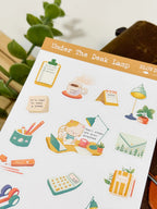 An angled close-up photo of a sticker sheet titled "Under The Desk Lamp." The cozy stationery-themed stickers show a cat writing at a desk next to a lamp with the text "Small steps are also progress," alongside a calculator, a mug of coffee, a calendar, a clipboard to-do list, and sticky notes.