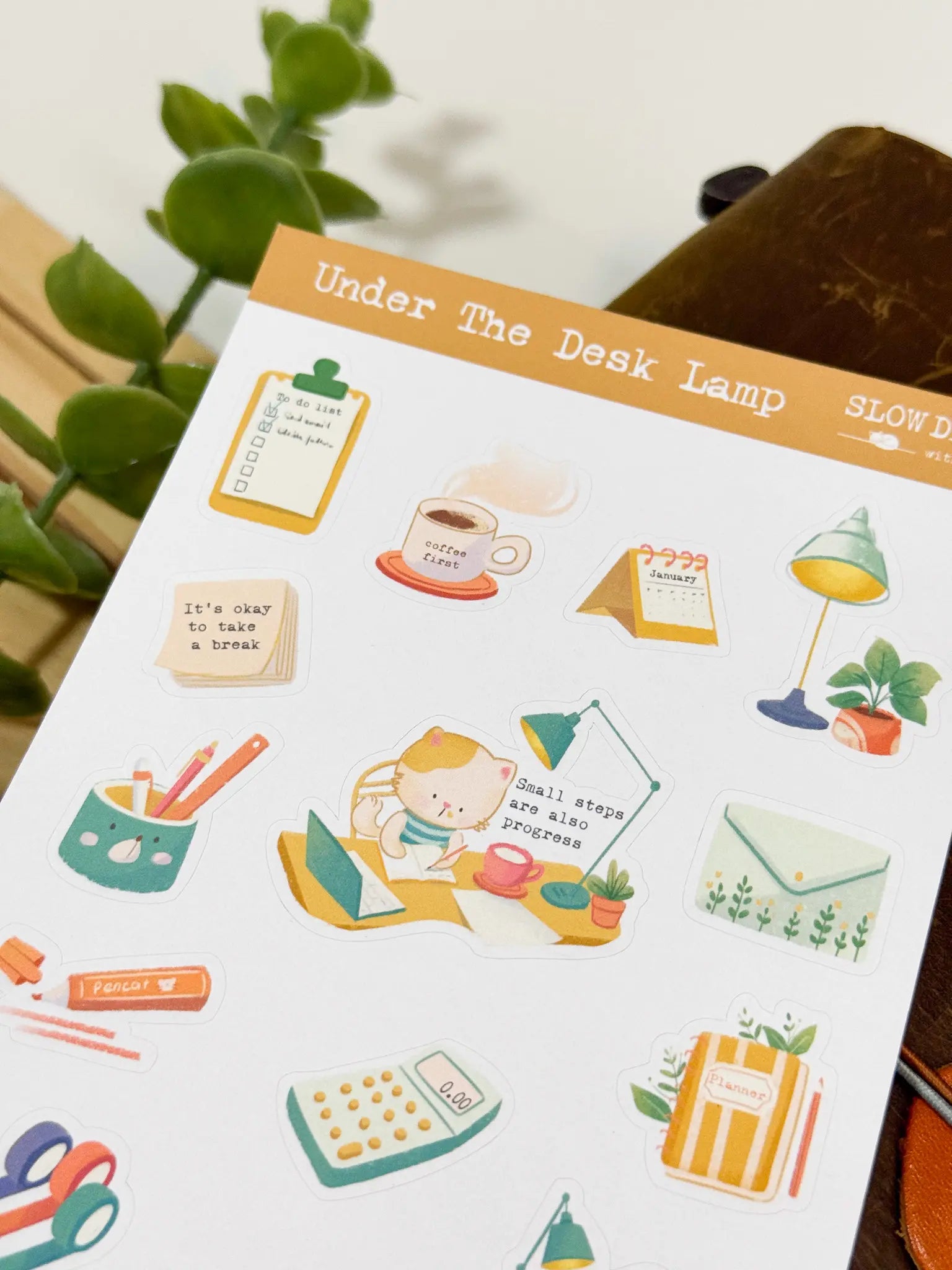 An angled close-up photo of a sticker sheet titled "Under The Desk Lamp." The cozy stationery-themed stickers show a cat writing at a desk next to a lamp with the text "Small steps are also progress," alongside a calculator, a mug of coffee, a calendar, a clipboard to-do list, and sticky notes.