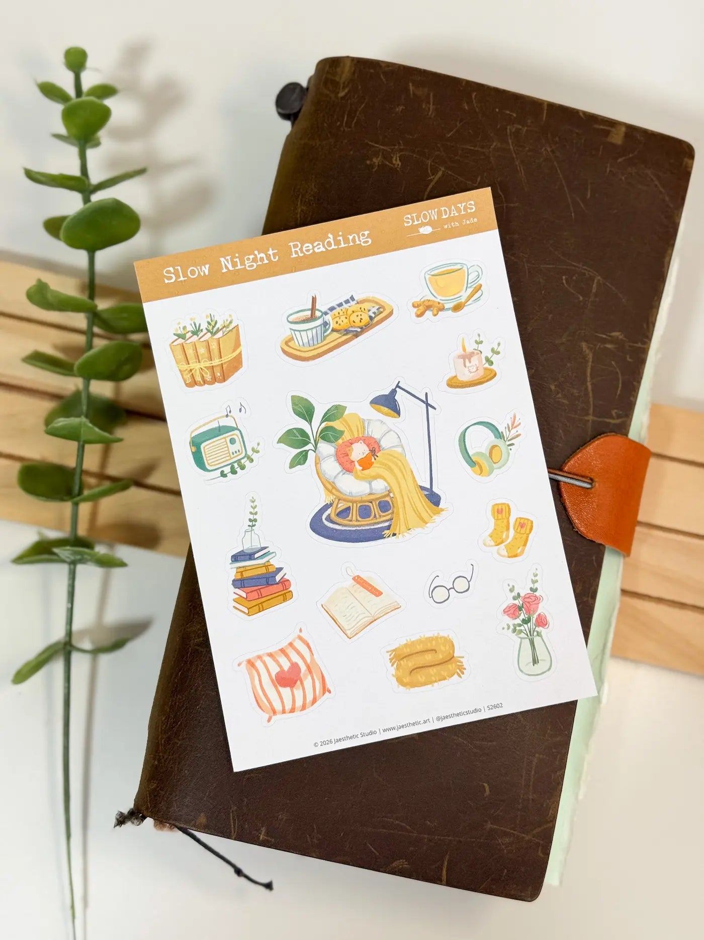 A full lifestyle flat lay photo of the "Slow Night Reading" sticker sheet styled on top of a rustic brown leather journal. A green leafy branch runs down the left background, beautifully showcasing the entire layout of cozy nighttime reading stickers.