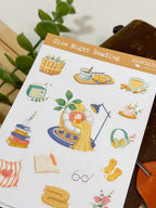 An angled close-up photo of a sticker sheet titled "Slow Night Reading." The stickers feature a cozy night-in theme, showcasing a cat tucked under a yellow blanket reading a book in a papasan chair under a lamp, stacks of books, a vintage radio, a steaming mug, and a pair of glasses.