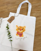 A white canvas tote bag laid flat on a wooden floor, featuring a whimsical watercolor illustration of an orange tabby cat wearing a red and white striped winter scarf. Beneath the drawing, the handwritten phrase "love at first sight" is written with a small heart. A brown kraft paper product tag is tied to the handle, and a sprig of green leaves lies beside the bag.