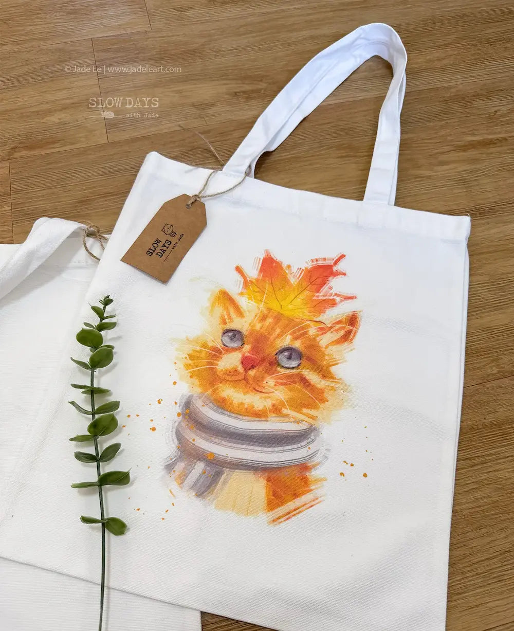 A white canvas tote bag laid flat on a wooden floor, featuring an autumnal watercolor illustration of a fluffy orange tabby cat wearing a gray and white striped scarf with a fallen maple leaf resting on its head. A brown kraft paper product tag is tied to the handle, and a sprig of green leaves lies beside the bag.