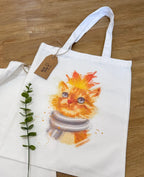 A white canvas tote bag laid flat on a wooden floor, featuring an autumnal watercolor illustration of a fluffy orange tabby cat wearing a gray and white striped scarf with a fallen maple leaf resting on its head. A brown kraft paper product tag is tied to the handle, and a sprig of green leaves lies beside the bag.