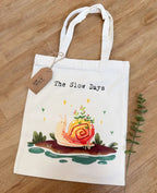 A white canvas tote bag laid flat on a wooden floor, featuring a whimsical illustration of a smiling pink garden snail with green plants growing from its pink and yellow shell. Above the illustration, it reads "The Slow Days" in a typewriter-style font, and a brown kraft paper product tag is tied to the handle "Slow Days With Jade"