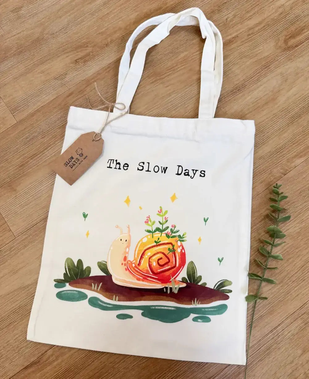 A white canvas tote bag laid flat on a wooden floor, featuring a whimsical illustration of a smiling pink garden snail with green plants growing from its pink and yellow shell. Above the illustration, it reads "The Slow Days" in a typewriter-style font, and a brown kraft paper product tag is tied to the handle "Slow Days With Jade"