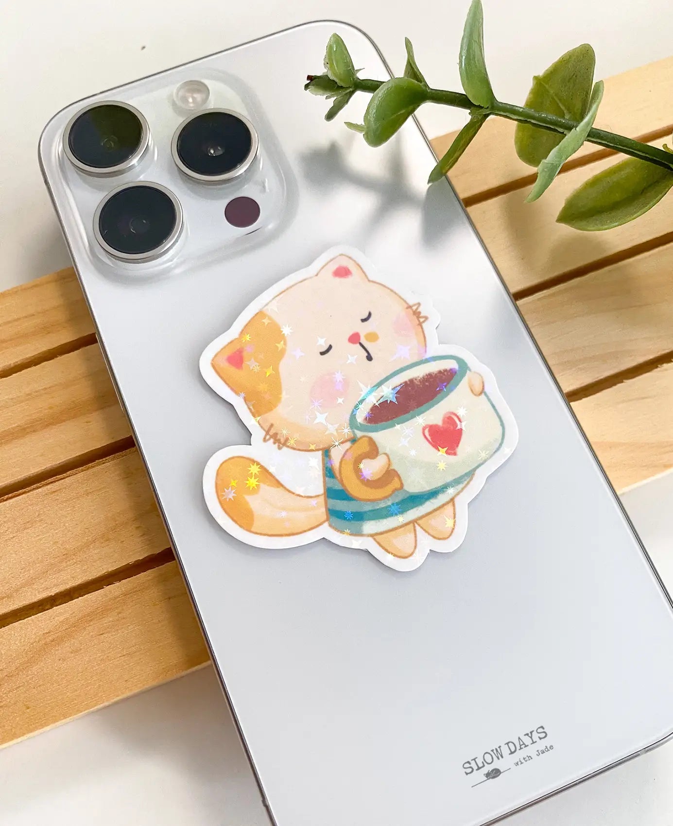 A lifestyle photo of the mug cat star holographic sticker placed on the back of a smartphone case to show its size, featuring an adorable cat hugging a mint green mug.
