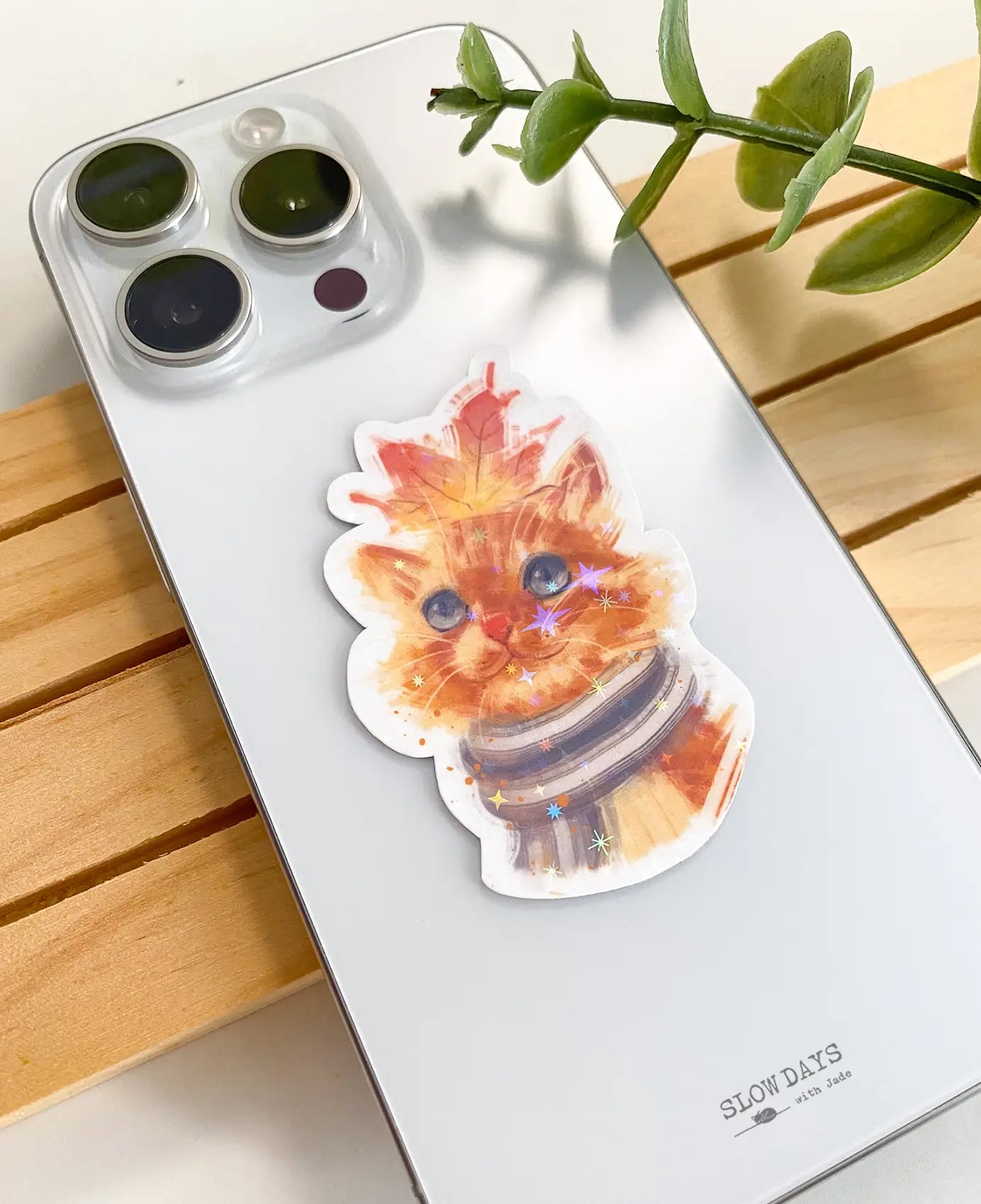 A lifestyle photo of the orange autumn leaf cat star holographic sticker placed on the back of a silver smartphone case to show its size. The sticker features an orange tabby cat wearing a striped scarf with a maple leaf on its head.