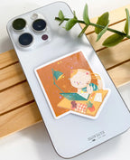 A lifestyle photo of the studying cat star holographic sticker placed on the back of a silver smartphone to show its size. The sticker shows a cat writing at a desk with a teal lamp, laptop, and plant on an orange background.
