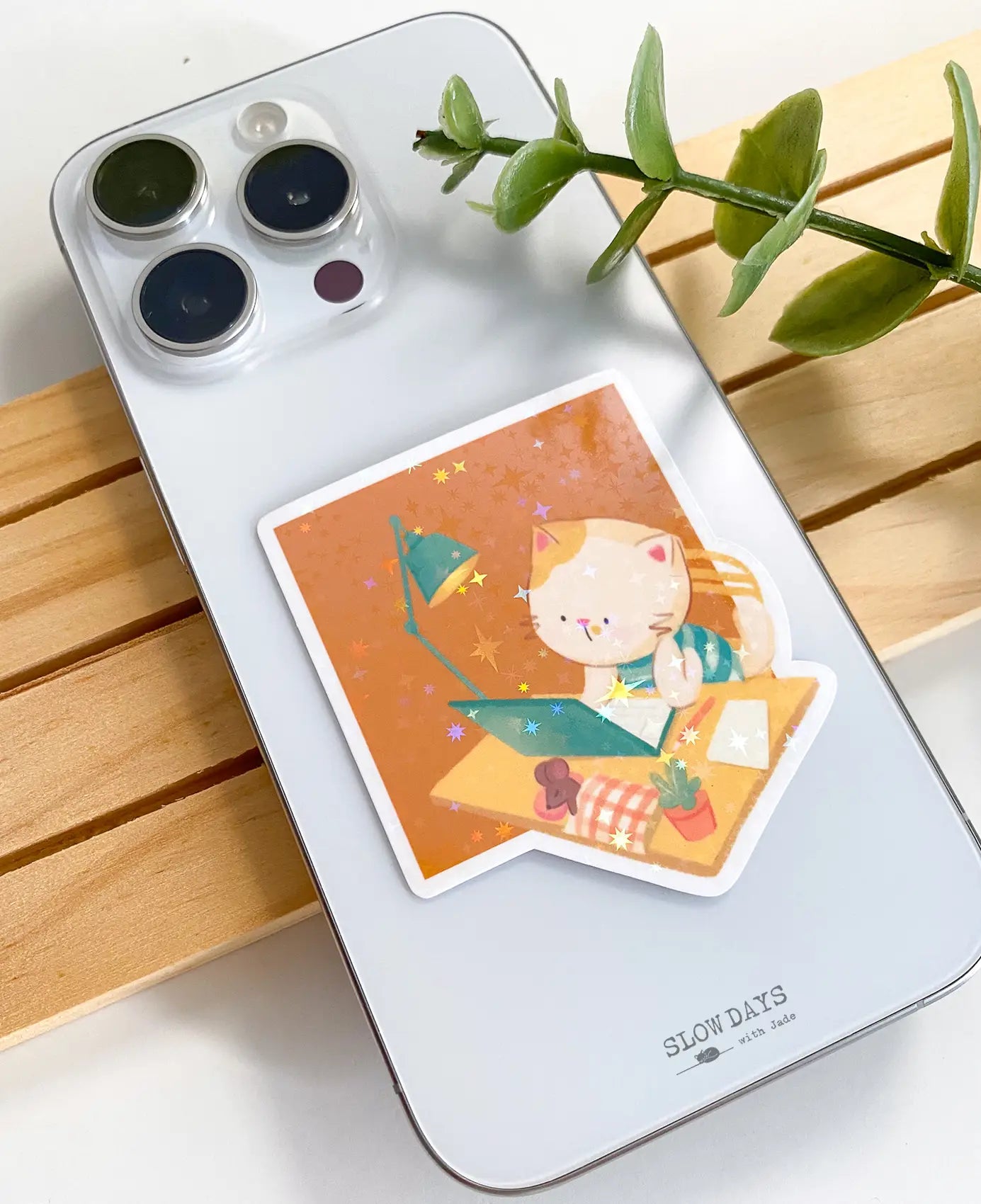 A lifestyle photo of the studying cat star holographic sticker placed on the back of a silver smartphone to show its size. The sticker shows a cat writing at a desk with a teal lamp, laptop, and plant on an orange background.