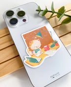 A lifestyle photo of a studying cat star holographic sticker placed on the back of a silver smartphone case to show its size. The sticker illustration features a cat sitting in a wooden chair writing in a notebook, next to a pink coffee mug, an open laptop, a teal desk lamp, and a potted plant against an orange background.