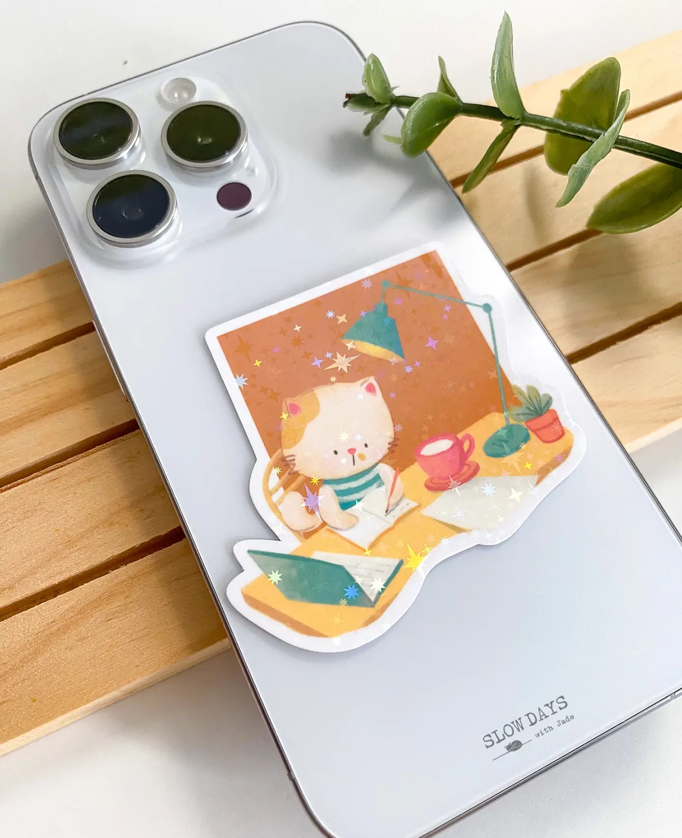 A lifestyle photo of a studying cat star holographic sticker placed on the back of a silver smartphone case to show its size. The sticker illustration features a cat sitting in a wooden chair writing in a notebook, next to a pink coffee mug, an open laptop, a teal desk lamp, and a potted plant against an orange background.