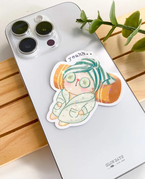 A lifestyle photo of the spa cat star holographic sticker placed on the back of a silver smartphone case to show its size. The sticker shows a relaxed cat in a mint green bathrobe and matching hair towel with cucumber slices over its eyes.