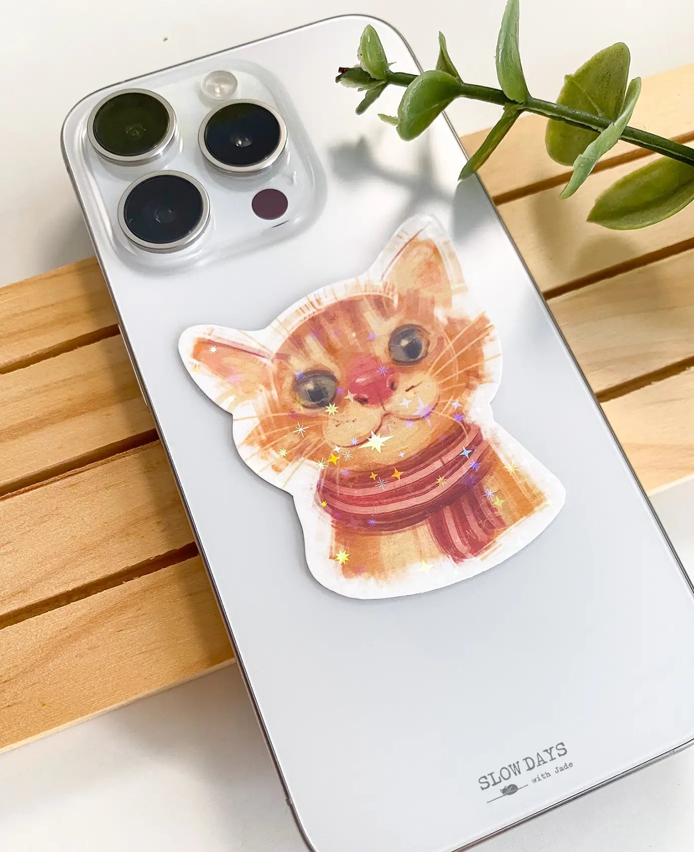 A lifestyle photo of the orange cat portrait star holographic sticker placed on the back of a silver smartphone case to show its size. The sticker shows an illustration of an orange tabby cat wearing a red and brown striped scarf.