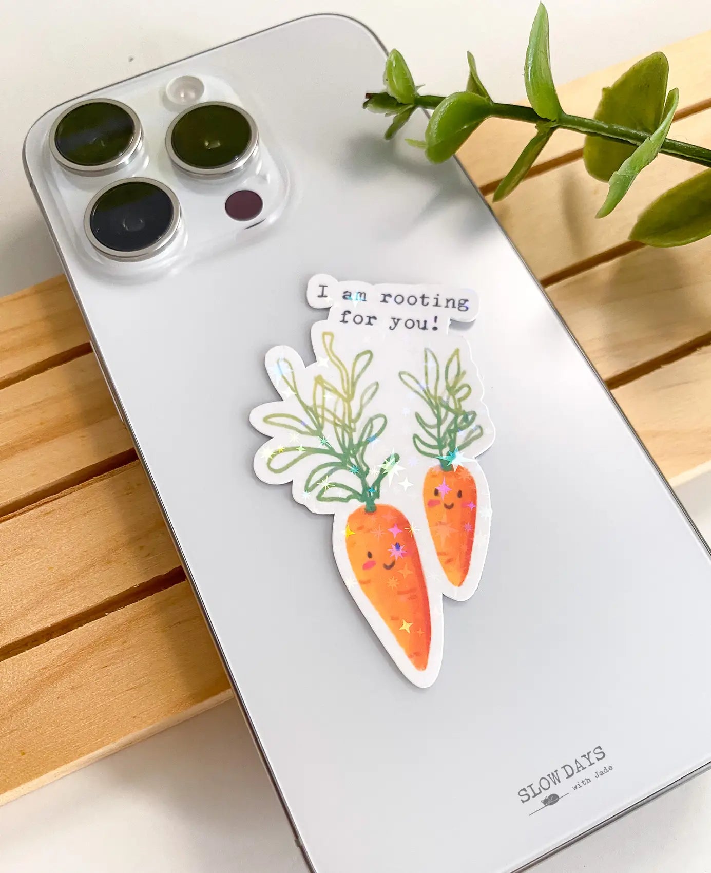 A lifestyle photo of the carrots quote star holographic sticker placed on the back of a silver smartphone case to show its size. The sticker features two smiling carrots with the text, "I am rooting for you!"