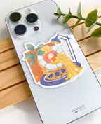A die-cut waterproof vinyl sticker featuring a cozy scene of a small cat tucked into a round papasan chair, reading an open book under a long yellow blanket. The background includes a large green potted plant and a dark blue floor lamp, all finished with a star holographic sparkle overlay. The sticker is placed on an iPhone.