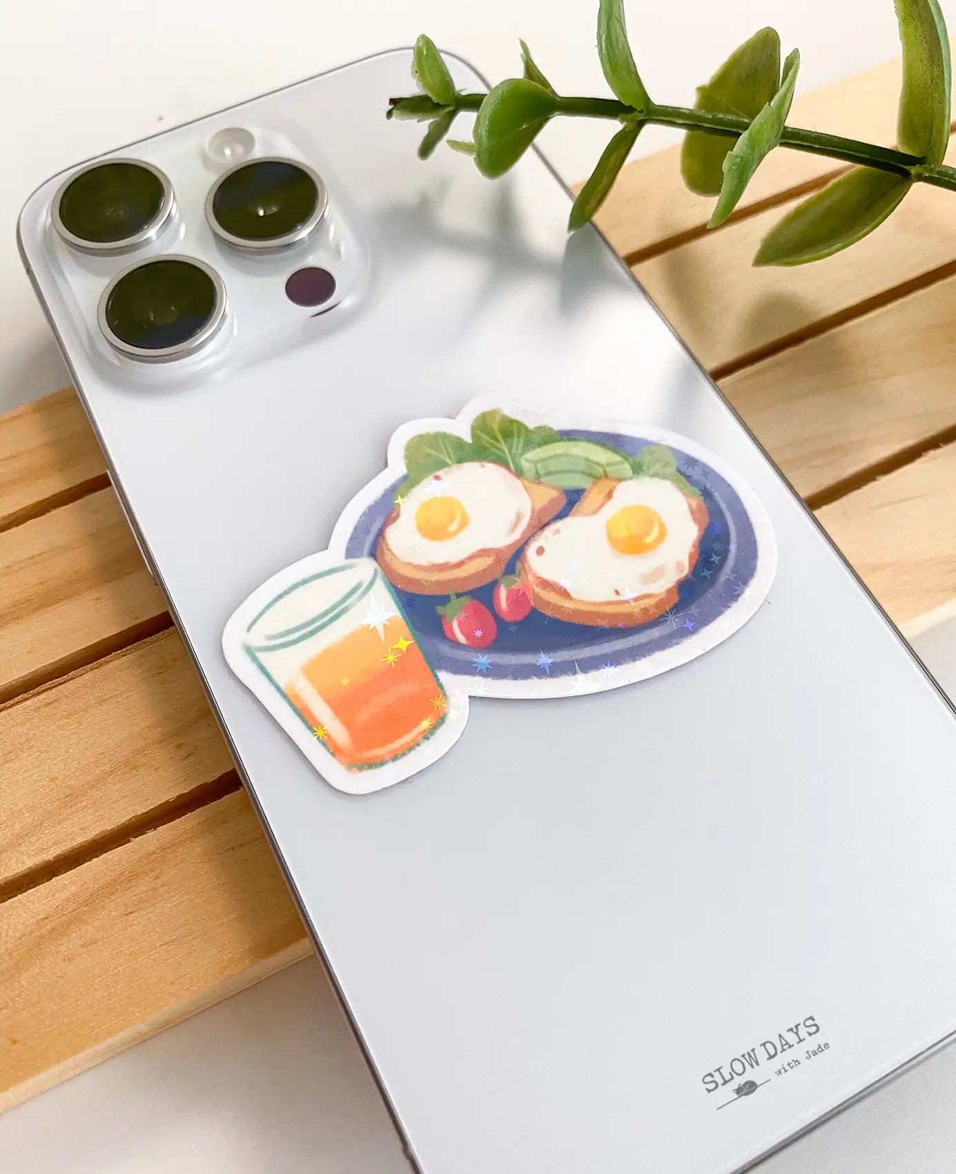 A lifestyle photo of the breakfast plate star holographic sticker placed on the back of a silver smartphone case to show its size. The sticker displays a blue plate with two sunny-side-up eggs on toast, avocado slices, and a glass of orange juice.