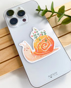 A lifestyle photo of the cute snail star holographic sticker placed on the back of a silver smartphone case to show its size. The sticker features a smiling pinkish snail with green plants and pink flowers growing out of its orange shell.