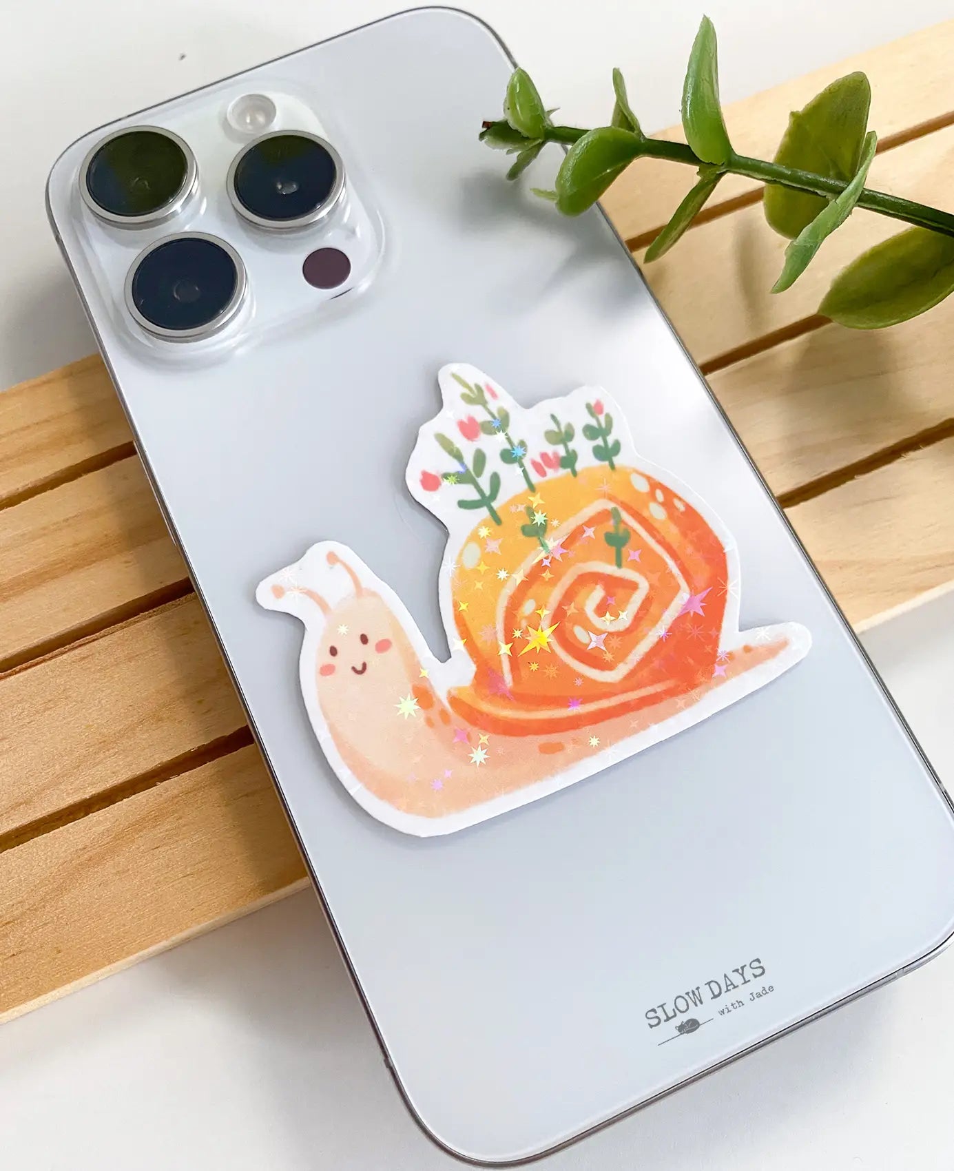 A lifestyle photo of the cute snail star holographic sticker placed on the back of a silver smartphone case to show its size. The sticker features a smiling pinkish snail with green plants and pink flowers growing out of its orange shell.