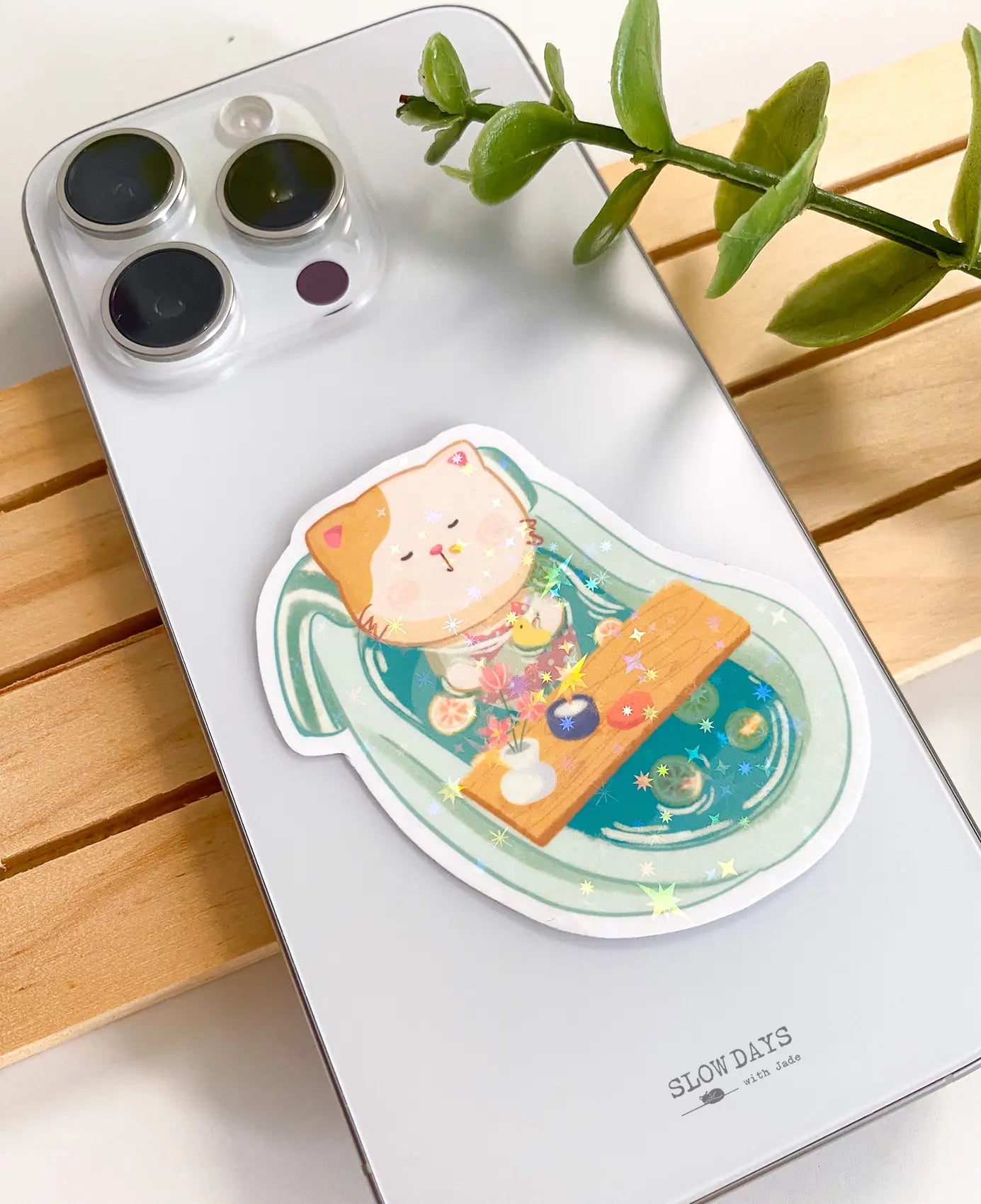 A lifestyle photo of the bathtub cat star holographic sticker placed on the back of a white smartphone case to show its size. The sticker features a cute cat relaxing in a mint green clawfoot bathtub.
