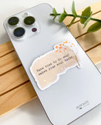 A lifestyle photo of the soul happy quote star holographic sticker placed on the back of a silver smartphone case to show its size. The textured beige sticker features a delicate branch and reads, "Take time to do what makes your soul happy."