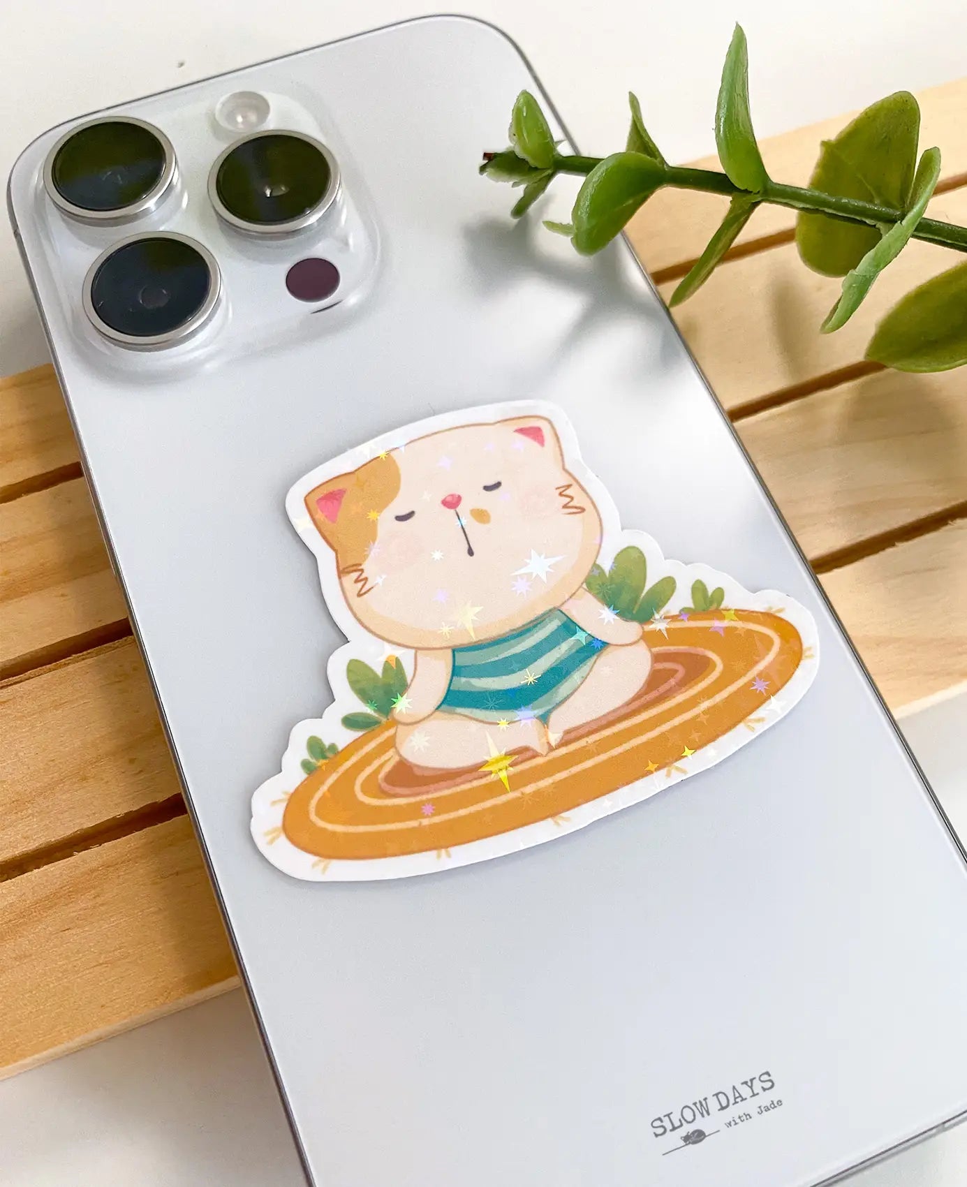 A lifestyle photo of the meditating cat star holographic sticker placed on the back of a smartphone case to show its size, featuring a cute cat on an orange striped rug.
